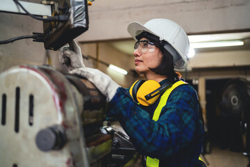 A woman in a blue plaid shirt and yellow safety gear is working on a machine. She is wearing a hard hat and safety glasses