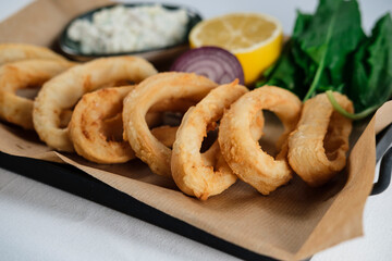 crispy fried calamari rings , golden brown crusty finger food served with dip sauce, greens and lemon, styled on plate with cooking paper