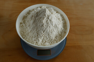 A bowl filled with a large quantity of white flour placed on a digital kitchen scale. The wooden table background adds a warm and rustic kitchen ambiance.  