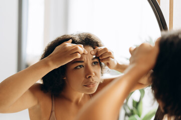 Young woman squeezing pimple on forehead in bathroom mirror