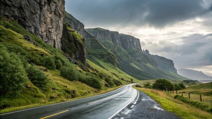 Naklejka premium Wet asphalt road winding through a picturesque valley, flanked by towering cliffs and lush green slopes under a cloudy sky, creating a dramatic and scenic landscape in iceland