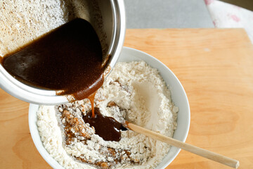 Making gingerbread. A bowl of flour and dry ingredients being mixed with dark liquid ingredients poured from a saucepan, showcasing the preparation stage of baking. 