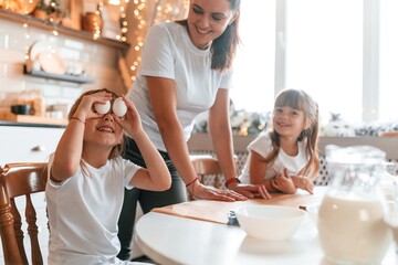 Holding eggs by the eyes. Mother with her little two daughters is preparing food on the kitchen with garlands together