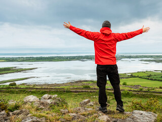 A man in a red jacket stands on a hill overlooking a valley and Galway bay. Male hands up in the air. Cloudy sky. Irish nature scene. Concept of travel and tourism. Burren, Ireland.