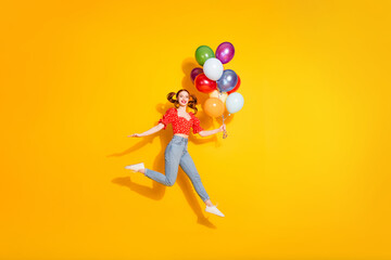 Photo of young funky positive girl wearing blouse and jeans jump trampoline with gifts air balloons isolated on yellow color background