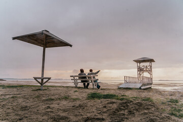 Backview of two adult guys, sitts on the bench and spending time by look around on the beauty of lake