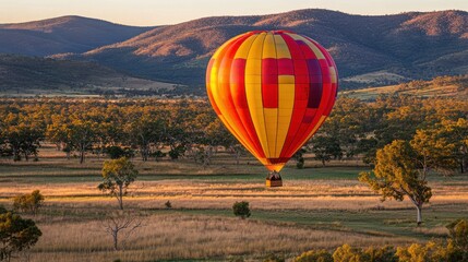 Obraz premium Colorful Hot Air Balloon Soaring High Above a Scenic Landscape at Sunrise with Lush Green Trees and Rolling Hills in the Background