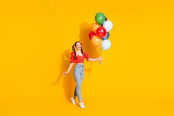 Full size photo of pretty young girl hold air balloons walking wear trendy red outfit isolated on yellow color background