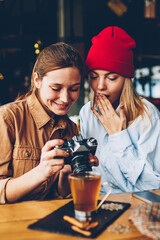 Wondered stylish teenager with cheerful friend female photographer dressed in casual outfit watching shocked photos on vintage camera during meeting in cafe interior enjoying recreation time