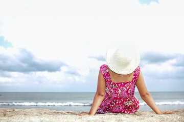 Young adult woman in pink floral beach dress sitting on beach sand under the sun with seascape and coastline on the background