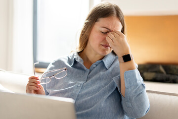 Tired woman working with laptop, sitting on sofa in living room at home