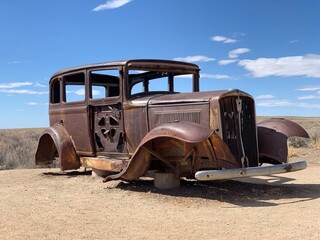 This image features a vintage rusted car, showing signs of significant wear and decay. The classic vehicle is positioned in an arid, desert-like landscape with a clear blue sky in the background.