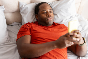 Young African American man reading news on mobile phone lying on bed