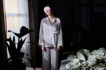 Sleepy woman dressed in crinkly cotton pajamas and pink sleep mask standing between bad and window...