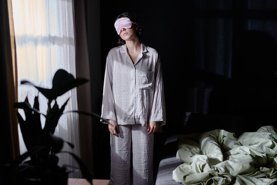 Sleepy woman dressed in crinkly cotton pajamas and pink sleep mask standing between bad and window illumed with moon light through window