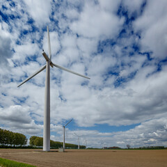 Several wind turbines stand in a plowed field, under a partly cloudy sky.  Rural landscape with trees.  Renewable energy.