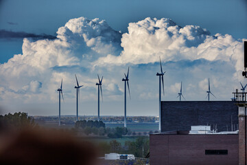 Wind turbines stand against a dramatic sky filled with large, puffy clouds.  A brick building and...