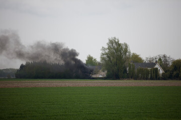 Dense dark smoke billows from a fire in a rural field, near a farmhouse.