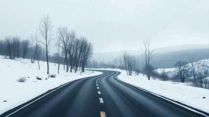 Obraz premium A winding asphalt road curves through a snow-covered landscape, bare trees lining the path under a muted winter sky.