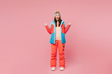 Full body skier young woman wear blue padded jacket ski goggles mask spend weekend winter season in mountains do winner gesture celebrate clench fists isolated on plain pink background. Hobby concept.