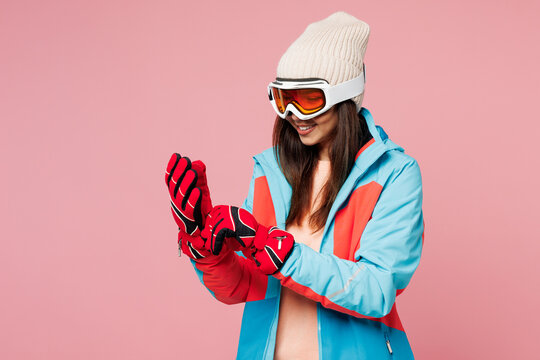 Side view snowboarder young fun woman wear warm blue padded jacket ski goggles mask spend extreme weekend winter season in mountains put on red glves isolated on plain pink background. Hobby concept.