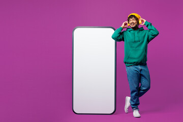 Full body young Indian man in green hoody hat casual clothes big huge blank screen area mobile cell phone smartphone listen music in headphones isolated on plain purple background. Lifestyle concept