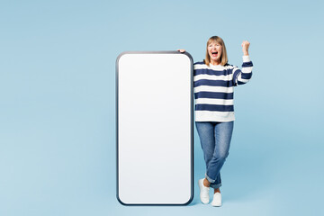 Full body elderly blonde woman 50s years old wears striped sweatshirt casual clothes big huge blank screen mobile cell phone smartphone with area do winner gesture isolated on plain blue background.