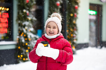 Cute little girl kid child with a paper cup of hot cocoa or tea in winter on the background of the snow city