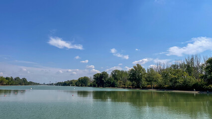 Wide view of a serene lake reflecting the sky, surrounded by lush trees under a bright blue sky, ideal for nature, relaxation, and eco-tourism themes.