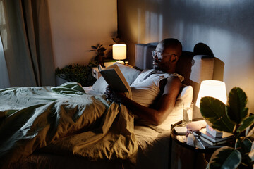 High angle shot of African American man enjoying reading book in comfortable bed before sleep