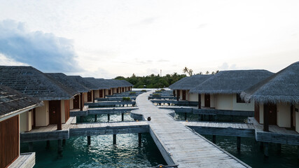 Bungalow on the water overlooking the green island. Thatched roofs, wooden bridge, water. Dark clouds. Green palm trees and a cell tower. A place to relax. Flowers grow in flower beds in places.