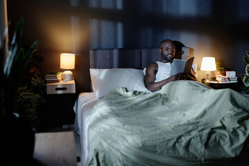 Mature African American man lying in comfortable bed and enjoying reading book at night in dark bedroom
