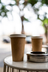 Brown coffee paper cup with a white paper lid and ashtray at outdoor table