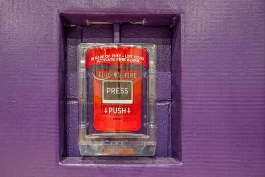 Red fire alarm system pull station under a protective clear plastic cover.  Fire alarm in a gymnasium mounted on purple wall with purple protective wall pads.