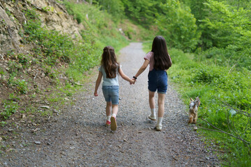 Two little girls walking hand in hand up a mountain path with a dog on a leash. Concept of sisterly friendship and family travel