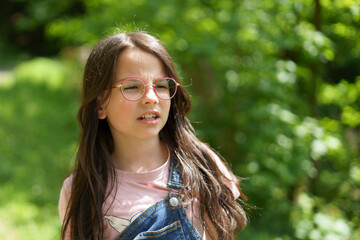 Little girl in glasses and denim overalls in summer outdoors in park. Concept of active healthy...