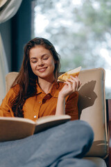 A happy young woman enjoying a healthy snack while reading a book in a cozy indoor setting, wearing a stylish amber shirt and relaxed denim