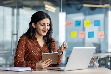 Confident woman in a workspace engages remotely on her laptop, jotting down important details while...