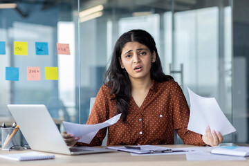 Businesswoman sitting at her desk while looking stressed amidst paperwork and a laptop.