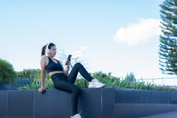 Young sportswoman wearing black sportswear and white headphones is sitting on a low wall using her smartphone in a city park, enjoying a break in her workout routine