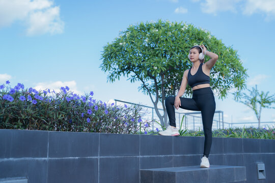 Young asian sportswoman wearing headphones and sportswear, resting after exercising outdoors in a park on a sunny day, taking a break from her workout routine