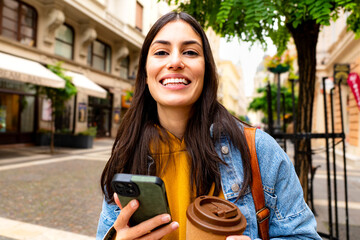 Young woman tourist smiling at the camera while holding a phone in one hand and a coffee cup in the...