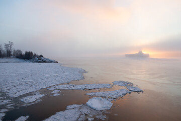 Cargo ship moving forward in front of the rising sun seen from a frozen river coast during a hazy winter dawn, Sainte-Pétronille, Island of Orleans, Quebec, Canada