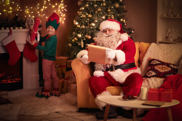 Santa Claus reading letter with cute little elf at home on Christmas eve