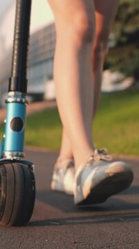 Close-up foot or leg of young woman walking on sidewalk with blue scooter. Girl walking in park on sunset. Teenager spends time alone at sunlight. Concept of college life, skating, summertime.