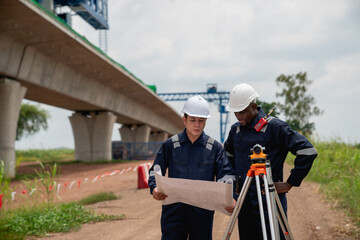 The survey team is using a theodolite and road construction plans to take measurements. Civil engineers are working with surveying tools to collect data for the construction project.