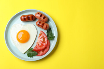 Plate of heart shaped fried egg and sausages on yellow background, top view. Space for text