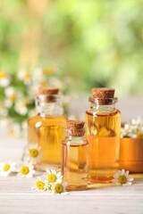 Bottles of essential oil and chamomile flowers on white wooden table against blurred background, closeup. Space for text