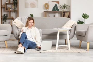 Fototapeta premium Young woman with cup of coffee using laptop on carpet at home