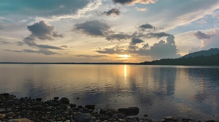 Fototapeta premium tranquil lakeside scene at sunset, with the sky filled with soft, majestic clouds and the lake reflecting the warm hues of the setting sun. The calm water and the rocky shoreline add to the peaceful 